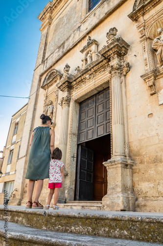 A mother and her child climb the steps and enter the church of Cavallino, Lecce, Puglia, Salento, Italy. In baroque style. Wooden portal with statues on the sides, onthe facade. © Ragemax