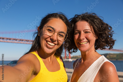 Happy female tourists enjoying walk and taking selfie outdoors. Two women standing on city promenade and smiling at phone camera. Happy tourists concept