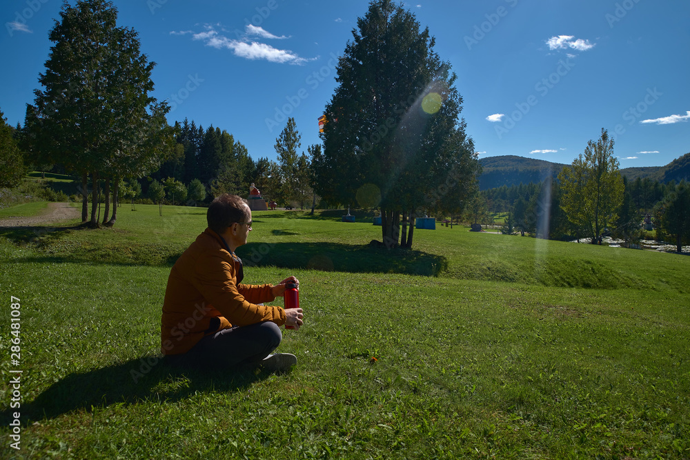 30-year-old man resting outdoors sitting on the grass on a camping trip holding a red flask water, in Quebec, Harrington. Travel to Canada 30-year-old man resting outdoors sitting on the grass on a camping trip holding a red flask water, in Quebec, Harrington. Travel to Canada