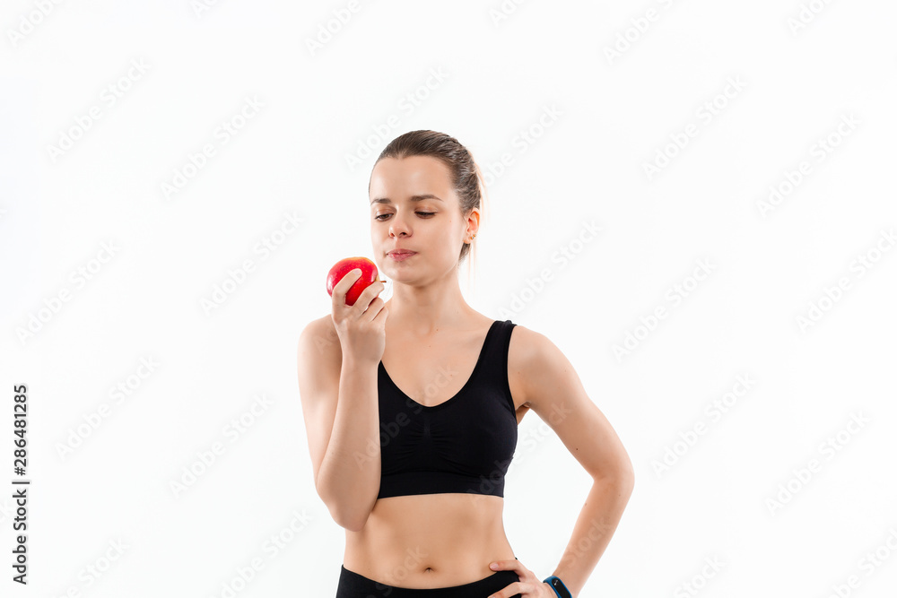 Young blond woman in a black sportswear with smart watches holding red apple after workout standing over white background. Young blond woman in a black sportswear with smart watches holding red apple after workout standing over white background.