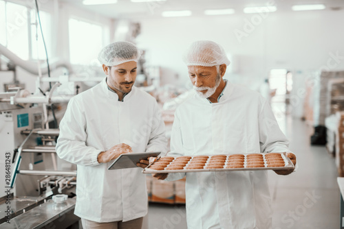 Senior adult employee holding tray with fresh cookies while supervisor evaluating quality and holding tablet. Both are dressed in sterile white uniforms and having hairnets. Food plant interior.