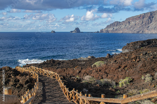 Volcanic pathway with wooden railing, with Roques de Salmor, Atlantic ocean, blue sky and clouds background,  in Charco de los Sargos, Frontera, El Hierro island, Canary islands, Spain photo