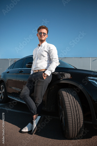 Young successful businessman in a white shirt and sun glasses standing near his luxary black car. © ianachyrva