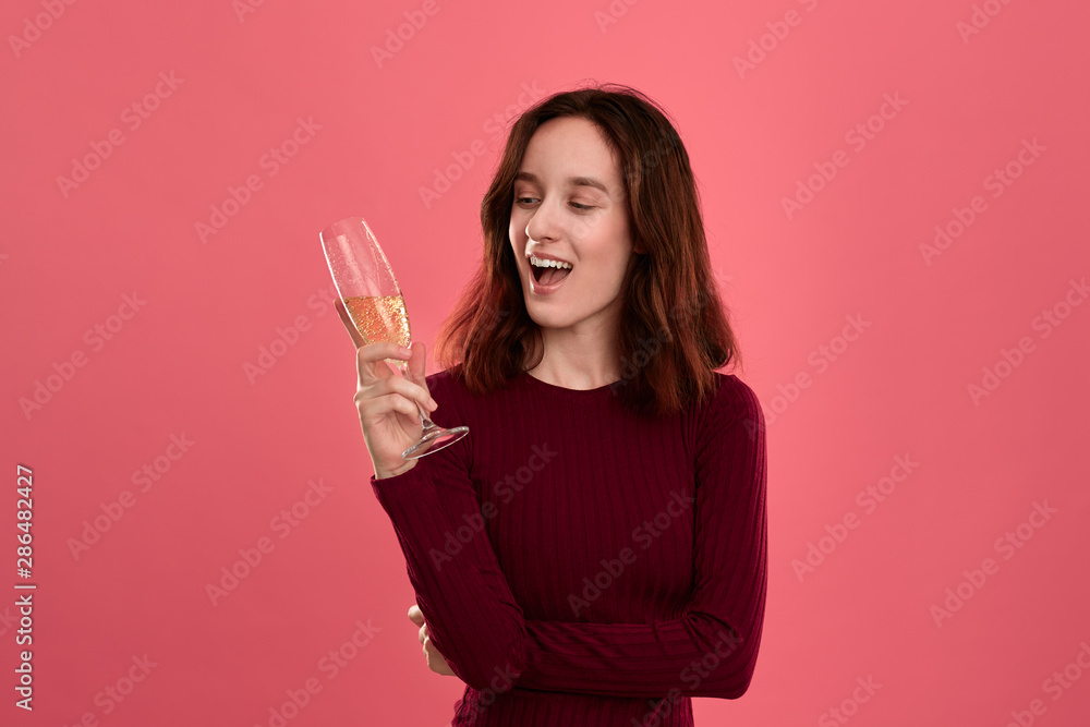 Excited pretty brunette girl holding wineglass of sparkling champagne standing isolated on a dark pink background and smiling at the camera. Excited pretty brunette girl holding wineglass of sparkling champagne standing isolated on a dark pink background and smiling at the camera.