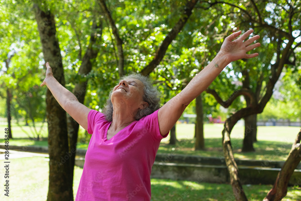 Happy carefree old lady exercising outdoors. Senior grey haired woman in casual with closed eyes standing in park, raising hands and looking up. Leisure outdoors concept Happy carefree old lady exercising outdoors. Senior grey haired woman in casual with closed eyes standing in park, raising hands and looking up. Leisure outdoors concept