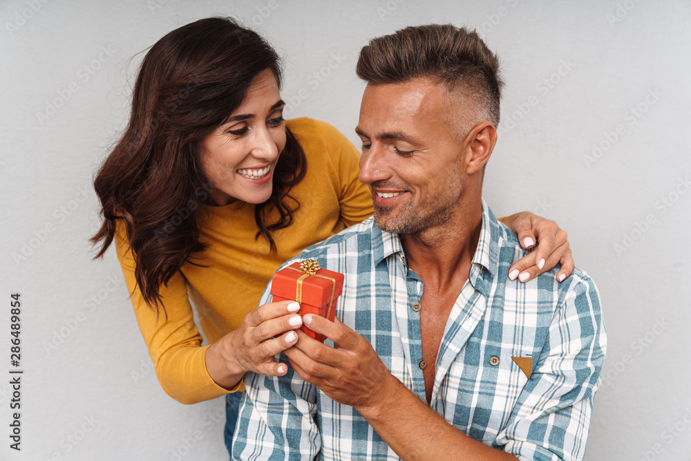 Happy cheerful woman give a present to her man isolated over grey wall background. Happy cheerful woman give a present to her man isolated over grey wall background.