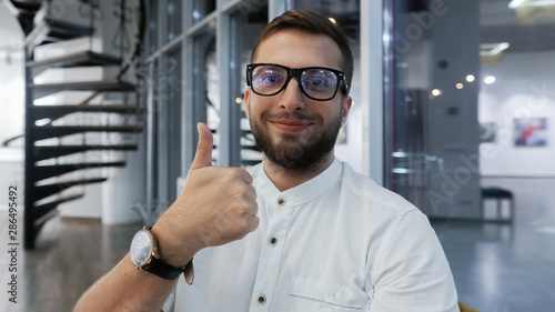 Portrait of a happy young man in eyeglasses and with a beard. Successful office worker holds thumb up and smiling at camera, on background, learning and education concept © Jimmy Tudeschi