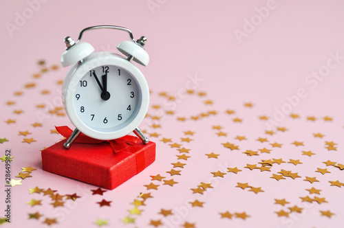 White alarm clock with red gift and star confetti on pink delicate background for holiday time.