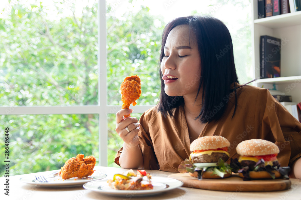 Chuby woman eating a large piece of fried chicken in the living room with a lot of fast food on the table. She stuck her tongue out of hunger and thirst and stared at the object with excitement.