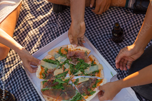 Top view of pizza in box and bottles of beer on picnic blanket. Man and women sitting on plaid, taking pizza slices and drinking beer. Pizza outdoors concept