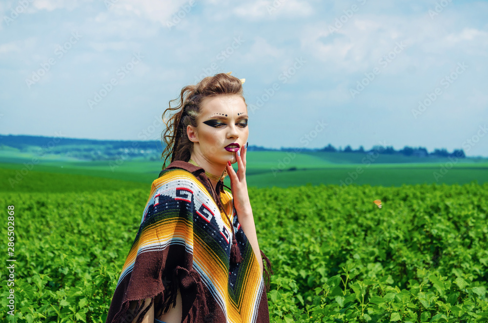 Foto Stock Portrait of a girl with makeup Amazon, Viking, aggressive war  paint. Dressed in a poncho on the background sky, green field | Adobe Stock