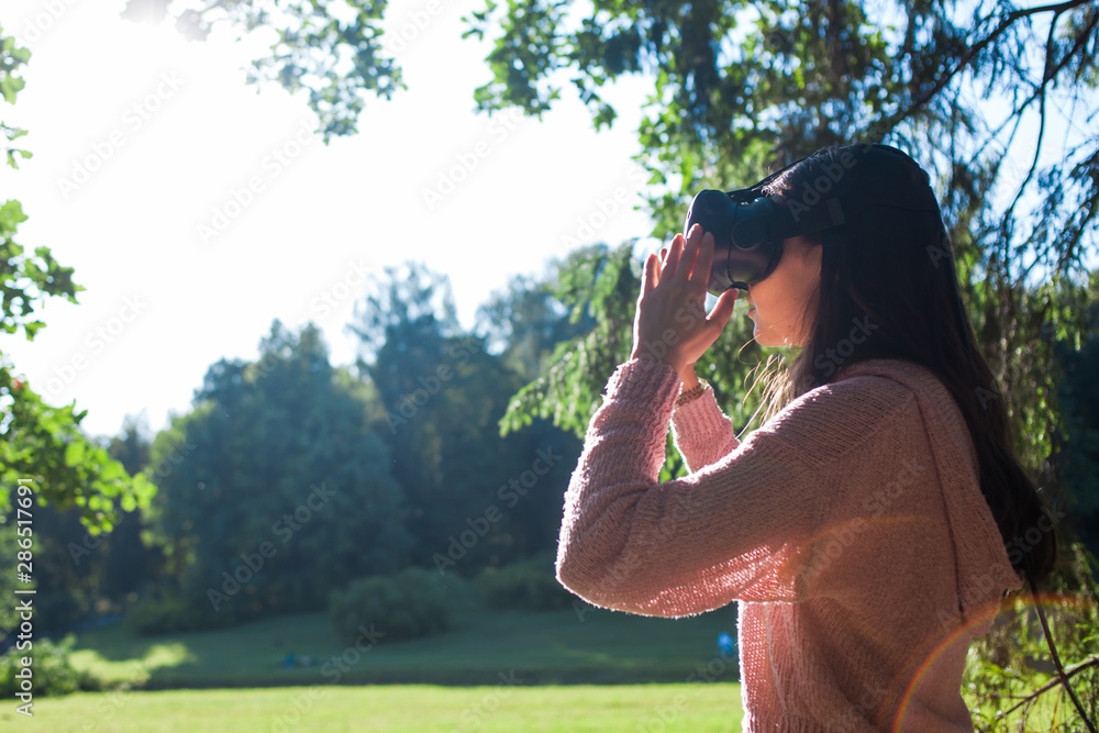Virtual forest. A young woman in VR glasses stands under the trees in a clearing, on sunny day, touches the air.