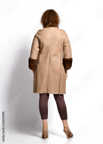 Tall woman with curly red hair stands back to us showing beige sheepskin coat with brown fur mandarin collar and cuffs