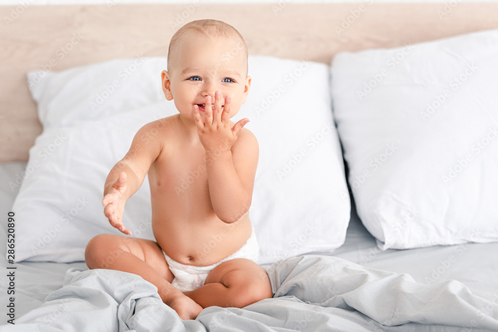 Adorable barefoot child in diaper smiling while sitting on white bedding Adorable barefoot child in diaper smiling while sitting on white bedding