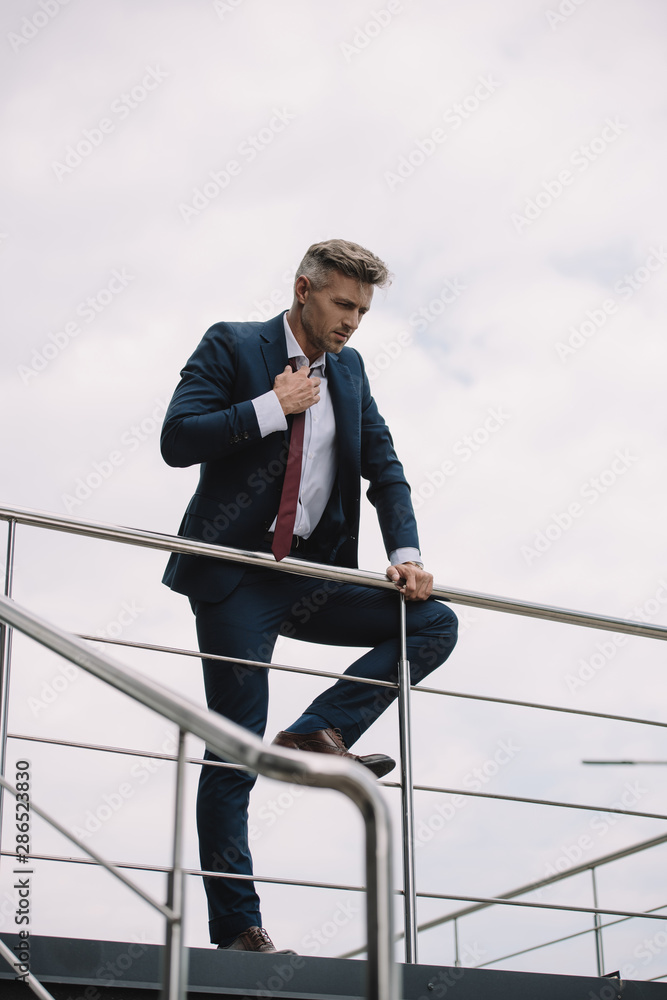 selective focus of upset businessman in suit standing outside and touching tie