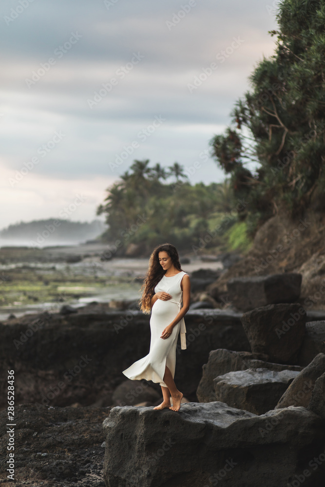 Young pregnant woman in white dress enjoying evening in Bali beach. Sensitivity to nature and harmony. Maternity concept.