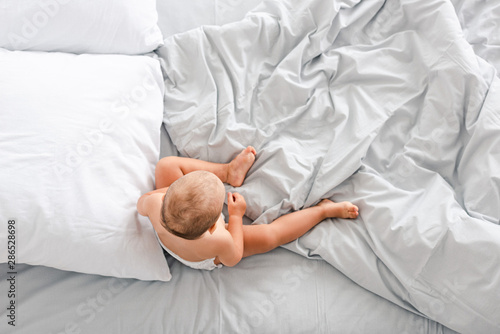 Top view of little child sitting on bed with crumpled light blanket