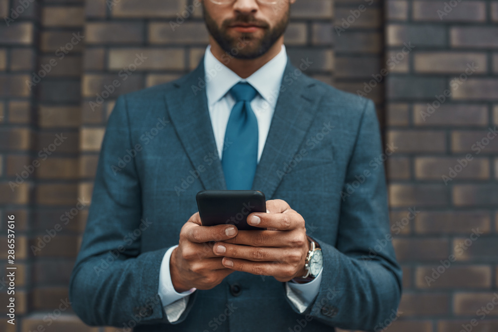 Cropped photo of a businessman in formal wear and using his smart phone while standing against brick wall Cropped photo of a businessman in formal wear and using his smart phone while standing against brick wall