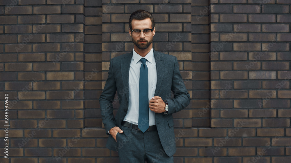 Great style. Confident businessman in full suit keeping hand in pocket and looking at camera while standing against brick wall Great style. Confident businessman in full suit keeping hand in pocket and looking at camera while standing against brick wall