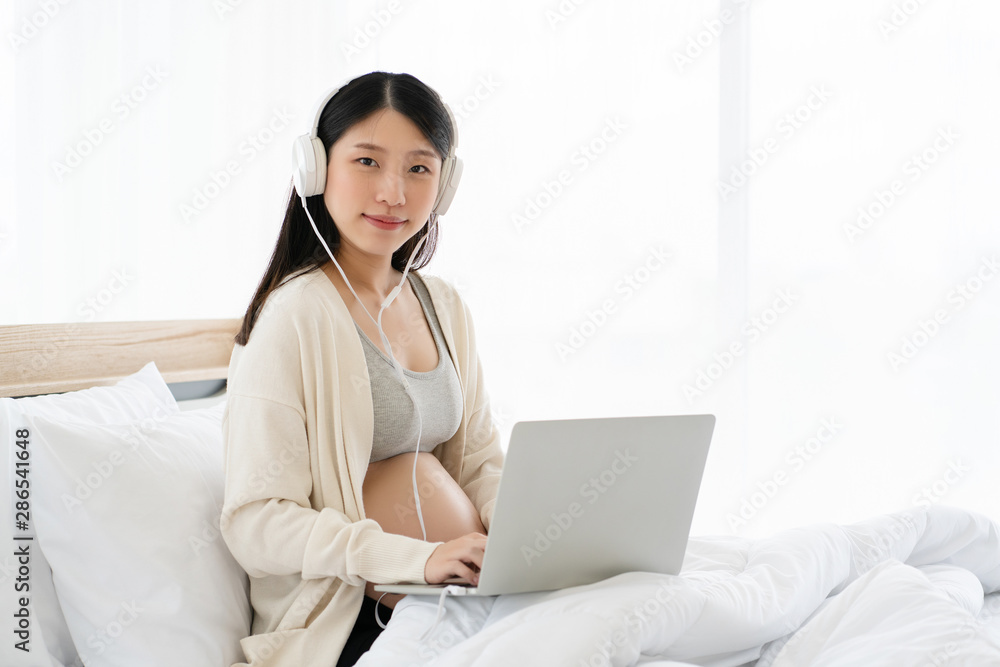 Pregnant Asian mother is using a laptop to shop online for newborn baby goods. In the bedroom And conducting export sales businesses online By being a sales blogger. Banner background with copy space Pregnant Asian mother is using a laptop to shop online for newborn baby goods. In the bedroom And conducting export sales businesses online By being a sales blogger. Banner background with copy space