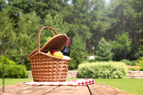 Fototapeta Naklejka Na Ścianę i Meble -  Picnic basket with fruits, bottle of wine and checkered blanket on wooden table in garden. Space for text