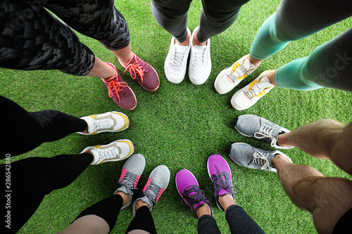 Closeup of athletes wearing sports shoes and standing in circle on green grass, top view. Space for text