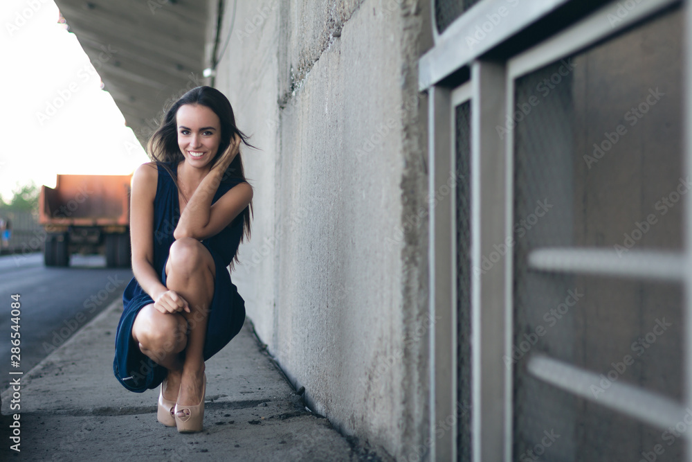 young elegant womana woman in a long dress squats on her heels against a long concrete wall stretching away into the distance. she smiles. cars on the background young elegant womana woman in a long dress squats on her heels against a long concrete wall stretching away into the distance. she smiles. cars on the background