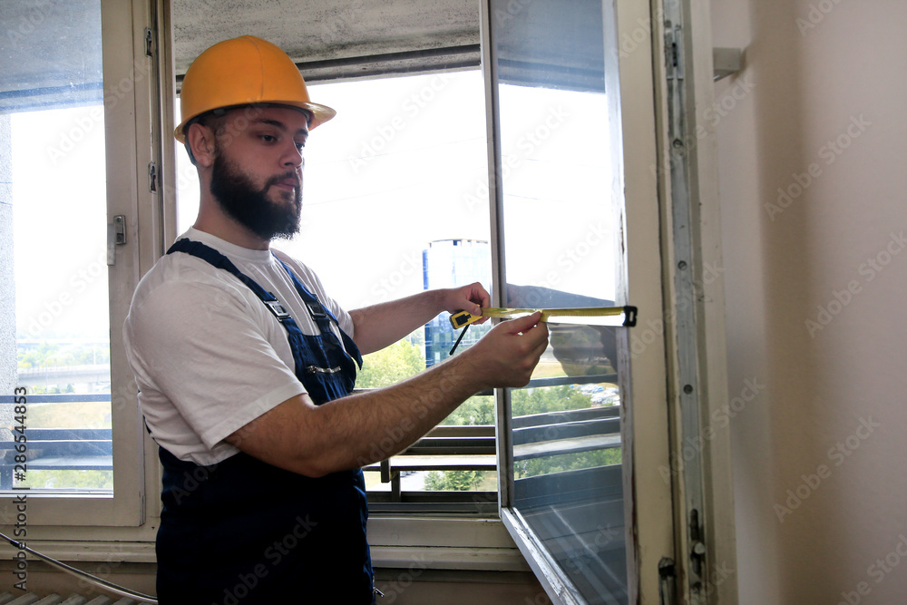 Construction worker and handyman is working on renovation of apartment. Builder is measuring window using measure tape at construction site. House renovation concept. Construction and service tools. Construction worker and handyman is working on renovation of apartment. Builder is measuring window using measure tape at construction site. House renovation concept. Construction and service tools.