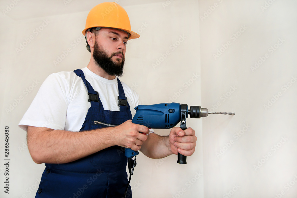 Construction worker and handyman works on renovation of apartment. Builder with blue electric drill drills a nail hole into wall of construction site. House renovation concept. Construction tools. Construction worker and handyman works on renovation of apartment. Builder with blue electric drill drills a nail hole into wall of construction site. House renovation concept. Construction tools.