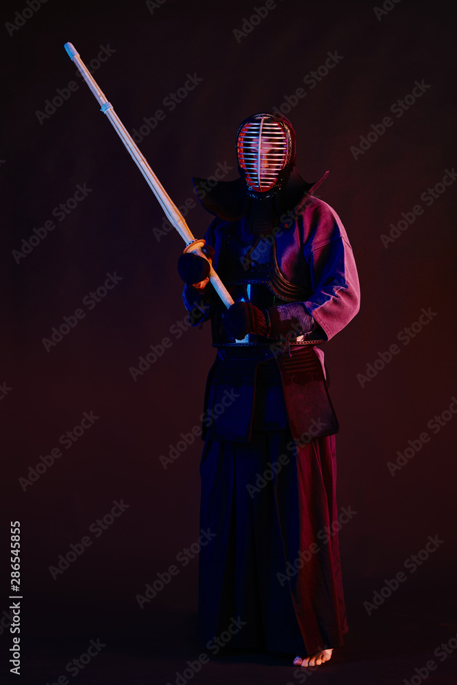 Fototapeta premium Bliska strzał, wojownik Kendo ubrany w zbroję, tradycyjne kimono, hełm uprawiający sztukę walki z bambusowym mieczem shinai, czarne tło.
