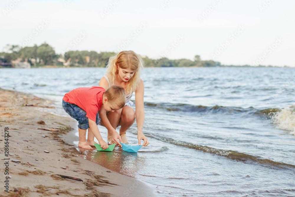 Naklejka premium Caucasian mother with toddler son child putting paper boats in water together on lake shore. Kid playing on beach. Outdoor family summer activity. Happy lifestyle childhood.
