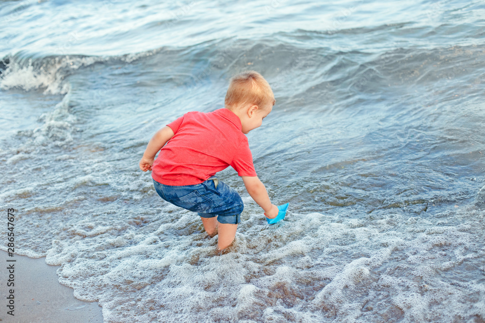 Happy Caucasian red-haired toddler child boy putting blue paper boat in water on lake sea ocean shore at evening sunset or morning. Kid playing on beach. Happy lifestyle childhood concept.