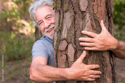 A senior adult man with white hair and beard hugging a tree in the wood - love for outdoors and nature - earth's day concept. People save the planet from deforestation