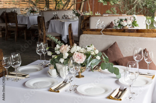Wedding table setting decorated with fresh flowers in a brass vase. Wedding floristry. Banquet table for guests outdoors with a view of green nature. Bouquet with roses, eustoma and eucalyptus leaves