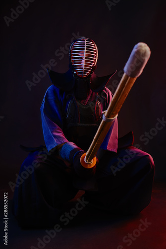 Close up. Kendo fighter wearing in an armor, traditional kimono, helmet, sitting, practicing martial art with shinai bamboo sword, black background.