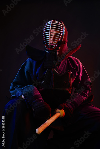 Close up. Kendo fighter wearing in an armor, traditional kimono, helmet, sitting, practicing martial art with shinai bamboo sword, black background.