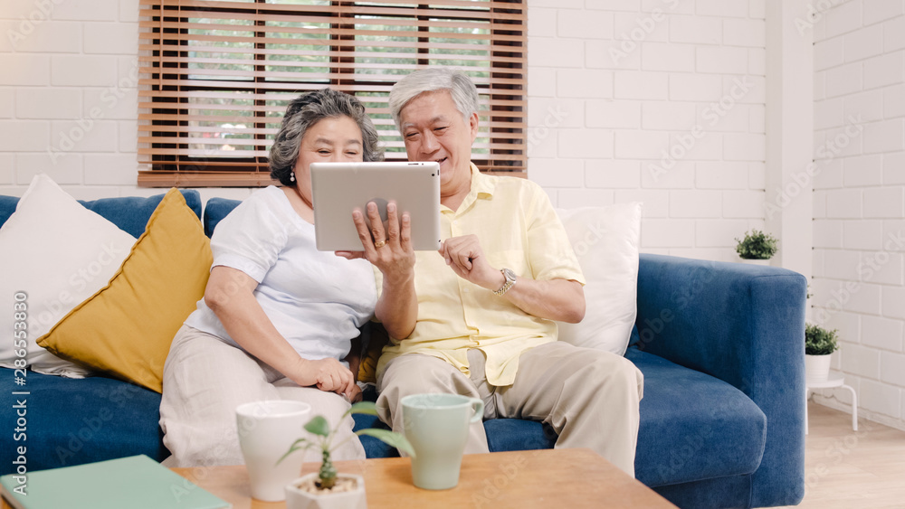 Asian elderly couple using tablet watching TV in living room at home, couple enjoy love moment while lying on sofa when relaxed at home. Enjoying time lifestyle senior family at home concept.