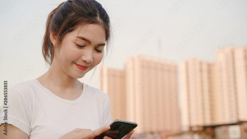 Cheerful Asian tourist blogger woman using touchscreen technology at smartphone while walking on the street at downtown city in the evening. Lifestyle backpack tourist travel holiday concept. Cheerful Asian tourist blogger woman using touchscreen technology at smartphone while walking on the street at downtown city in the evening. Lifestyle backpack tourist travel holiday concept.