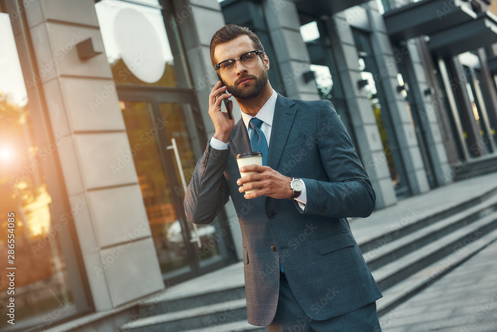 Important talk. Young and handsome bearded businessman in formal wear talking by phone and holding cup of coffee while standing outdoors Important talk. Young and handsome bearded businessman in formal wear talking by phone and holding cup of coffee while standing outdoors