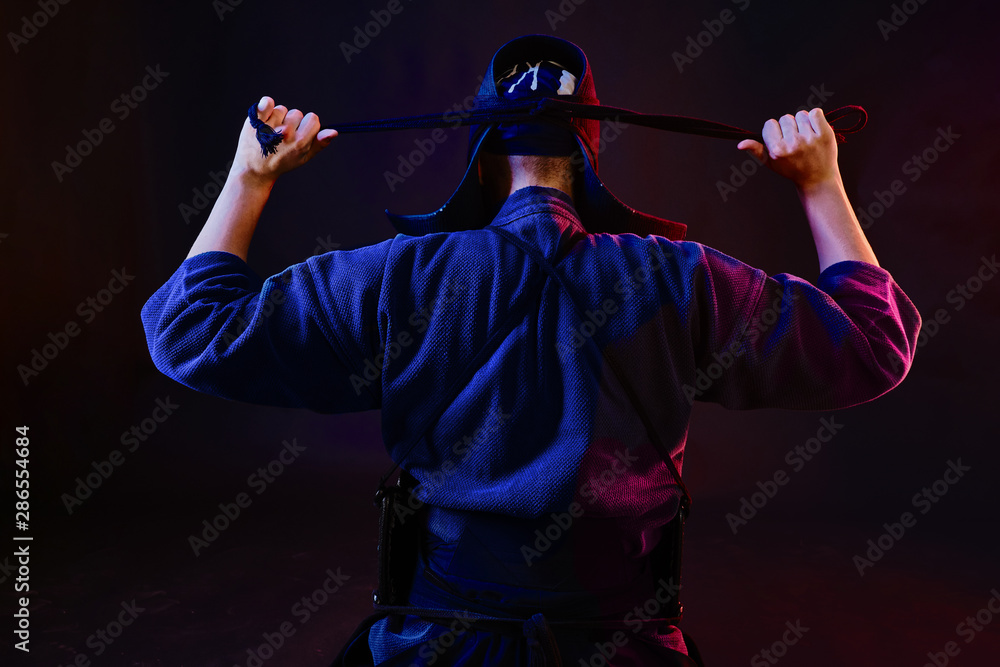 Close up shot, Kendo fighter wearing in an armor and traditional kimono is tying the lacing on his helmet standing back against a black background. Close up shot, Kendo fighter wearing in an armor and traditional kimono is tying the lacing on his helmet standing back against a black background.