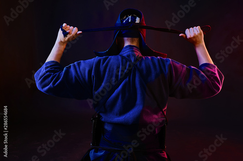 Close up shot, Kendo fighter wearing in an armor and traditional kimono is tying the lacing on his helmet standing back against a black background.