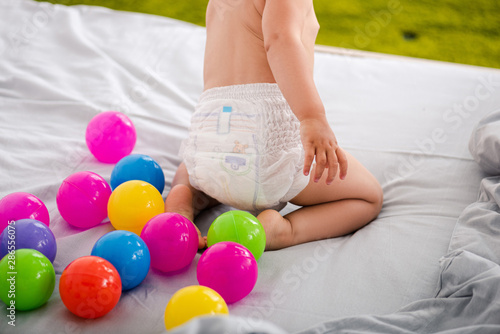 Cropped view of cute baby in diaper sitting on bed among colored balls