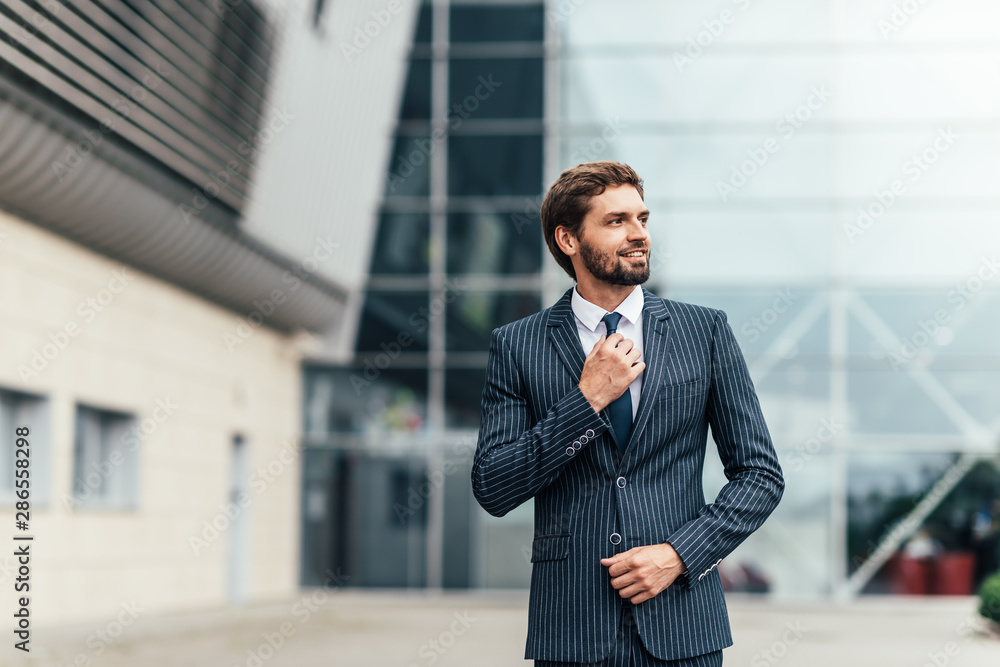 Confident businessman. Confident young man in full suit adjusting his sleeve and looking away while standing outdoors with cityscape in the background Confident businessman. Confident young man in full suit adjusting his sleeve and looking away while standing outdoors with cityscape in the background