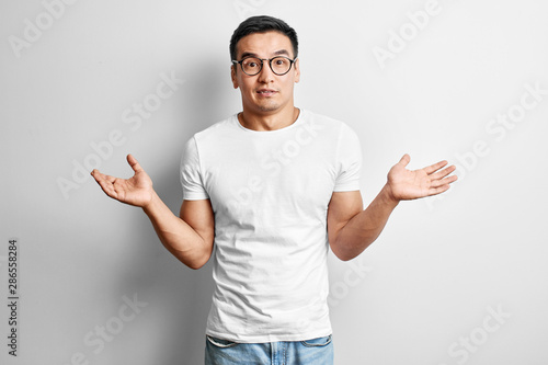 Puzzled Asian man dressed in casual and glasses with arms outstretched looks to the camera. Surprised, confused young Kazakh doesn't know what to do on white studio background