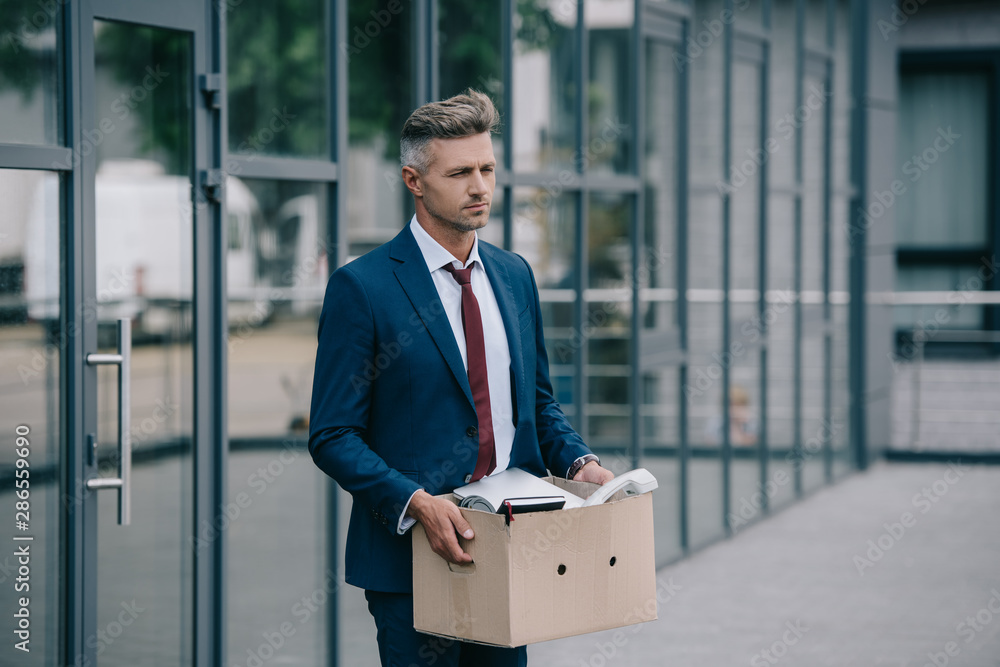 sad and fired businessman standing near building and holding carton box sad and fired businessman standing near building and holding carton box