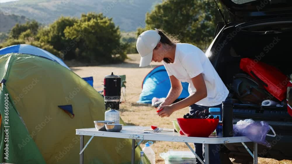 Young woman slicing tomato on the table near a tourist tent at camping. Female tourist cooking vegetable salad for breakfast in expedition camp Young woman slicing tomato on the table near a tourist tent at camping. Female tourist cooking vegetable salad for breakfast in expedition camp