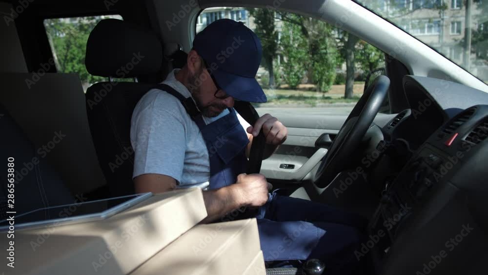 Adult worker of speed delivery service getting in car, fastening safety belt, starting and driving to deliver order. Delivery man in cap and glasses driving van, boxes and tablet pc lying next to him Adult worker of speed delivery service getting in car, fastening safety belt, starting and driving to deliver order. Delivery man in cap and glasses driving van, boxes and tablet pc lying next to him