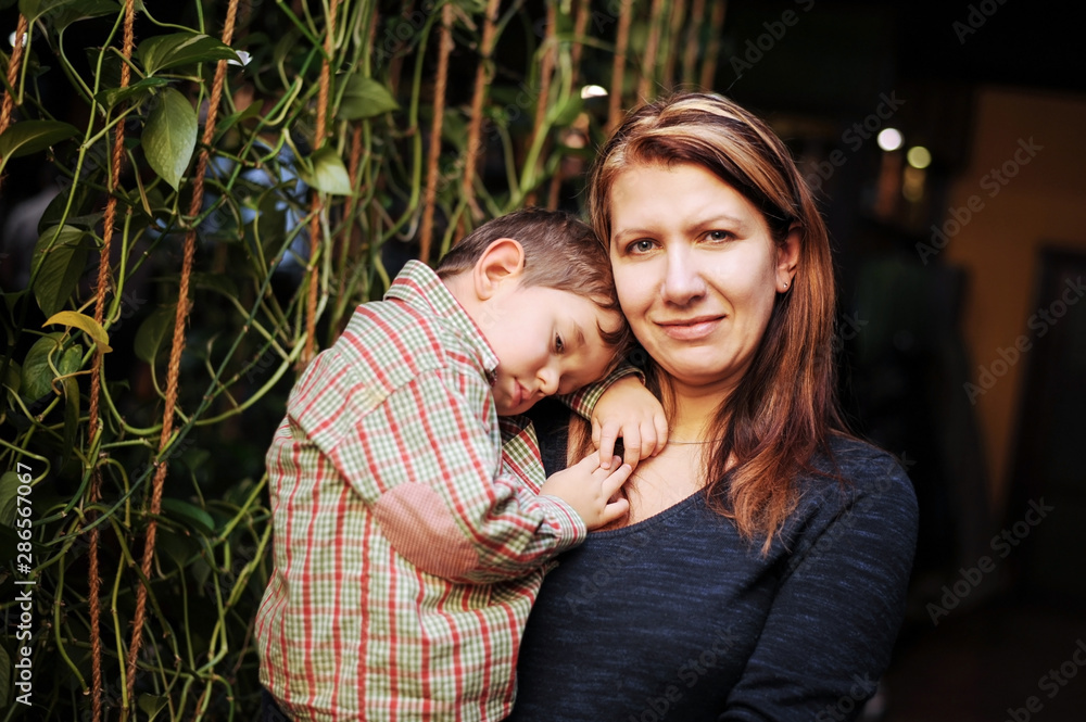 Fototapeta premium A woman holds in her hands a little boy who falls asleep in her arms. The boy rested with his mother in a cafe, had a good lunch and wanted to sleep