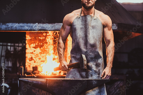 Canvas Print Muscular blacksmith in protective apron at his workshop with hammer in his hands