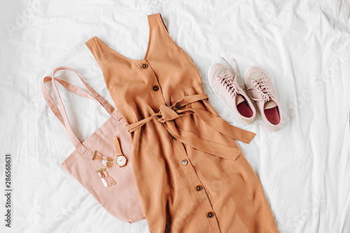 Beige dress with eco bag, sneakers and accessories on white bed. Women's stylish autumn or summer outfit. Trendy clothes. Flat lay, top view. photo